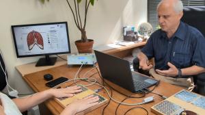 Doctor in blue shirt at desk explaining results to patient whose hands rest on Sensitiv Audit sensor plates; computer monitor shows lung diagram and Sensitiv Audit device on desk in clinic office.