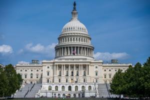 The grand West Front of the U.S. Capitol Building. The neoclassical white building, with its towering central dome and a statue on top, is centered against a clear blue sky with a few wispy clouds. An American flag is flying from a staff just below the do