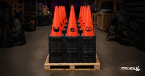 Stacks of orange Halloween traffic cones with black bases on a wooden pallet in a dim warehouse. The three front cones show different jack o' lantern faces, and Halloween props sit in the background.