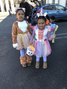 Two young girls stand in a parking lot wearing matching unicorn-themed costumes with ear headbands, one in a brown furry outfit with a white beard and the other in a pink and white ruffled dress. They hold a white bucket decorated with a panda face. Decor