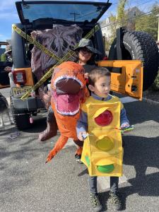 A young boy wearing a yellow traffic light costume with red, yellow, and green lights holds a lollipop and candy while standing in a parking lot. Behind him, a Jeep is decorated with caution tape and a large inflatable dinosaur head, with an adult in a di