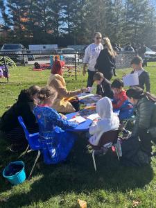 A group of children in costumes sit at a round outdoor table making crafts, with adults supervising nearby. A blue bucket sits on the grass in the foreground, with cars and a fence visible in the background.