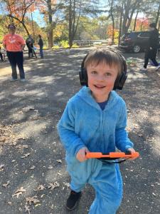 A young child wearing a light blue furry costume and black noise-canceling headphones smiles while walking down a paved path, holding an orange handheld device. Adults and a parked SUV are visible in the background among trees with autumn foliage.