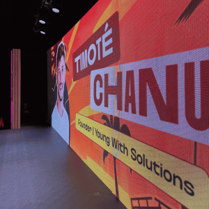 Large event screen displaying 'Timoté Chanut, Founder, Young With Solutions' with his photo, on stage at a speaking event.