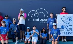 Group of Love Serving Autism participants, coaches and volunteers posing together in front of a Miami Open sign, holding a Love Serving Autism banner.