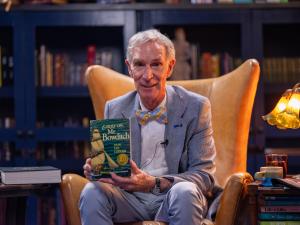Bill Nye poses in a high-backed leather library chair, holding his copy of "Carry On, Mr. Bowditch" by Jean Lee Latham.