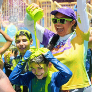 Woman in sunglasses pours water from a green cup over a smiling child at a colorful outdoor water festival.