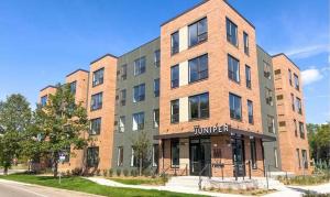 Exterior of The Juniper, a four-story affordable housing building with brick and gray metal panel siding, in Maplewood, Minnesota, with a covered entrance reading 'Juniper' and landscaped walkway.