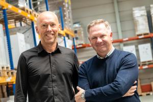 Hayco van Gaal en Noah Rogers staan naast elkaar in een magazijn bij de fabriek van RPI Print in Eindhoven, beiden glimlachend naar de camera. Van Gaal, links, draagt een zwart overhemd met knopen. Rogers, rechts, draagt een marineblauwe trui over een geruit overhemd