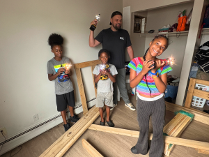 An SHP volunteer stands with three children holding illuminated DEWALT drills as they help assemble their own wooden bed frame during a power outage.