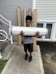 A young boy carries a rolled-up mattress outside a house, with wooden bed frame boards leaning against the wall behind him.
