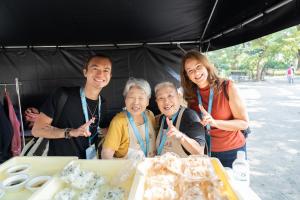 Two local grandmothers in aprons smile with two digital nomad participants behind a table of rice balls at Colive Fukuoka 2025.