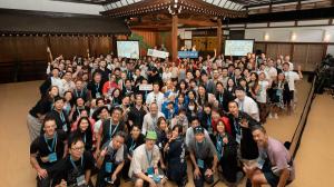 Group photo of Colive Fukuoka 2025 participants in a traditional Japanese hall, many wearing event lanyards and waving.