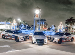 Three police vehicles, including two marked police cars and an SUV, parked in front of a festively decorated outdoor area with blue and white holiday lights at night.
