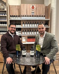 Cosku Kaplan and Umut Kaplan seated at Coccinella with Oleavia Havaalani I and South Aegean Special bottles and their 2026 NYIOOC Silver Award trophies