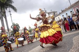 Women in yellow and red dresses and ornate headdresses dance and play drums in a cobblestone square lined with colonial buildings, in the city of Salvador Bahia, Brazil, while a crowd of people wearing event lanyards watches.