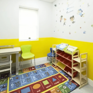 Colorful children’s playroom with educational rugs, orange chairs, activity wall, toy shelves, tables, and a green sofa.
