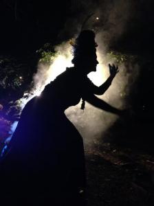 Silhouette of the Stick Witch, a costumed Victorian Ghost Walk character, reaching through backlit fog at night in Old Louisville's Central Park