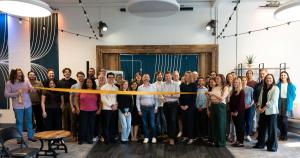 A large group of people stand together indoors, smiling, as two people in the center cut a wide yellow ribbon with scissors. The space has string lights, wooden accent walls, and a teal curtain backdrop.