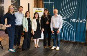 Six people stand smiling side by side indoors in front of a teal curtain with the Revive logo, dressed in business attire.