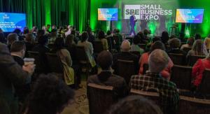Audience members face a green-lit stage displaying the Small Business Expo logo during a keynote presentation.