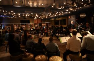 Actors seated in a circle at music stands perform The Weight of Silence beneath string lights and framed rodeo photographs