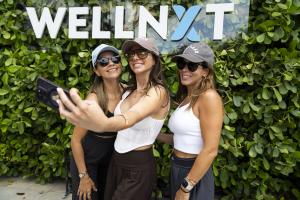 three women taking a selfie in front of a WellNXT sign with green boxwood behind it