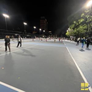 Crowd on the McCarren Park public tennis courts at night, lit by LED sports lights on tall poles along both sides of the courts.