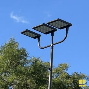 Three rectangular LED sports light fixtures mounted on a curved triple bracket atop a bronze pole against a blue sky.