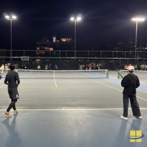 Two tennis players stand at the baseline of a lit court at night while a crowd gathers along the net and fence under LED sports lights.