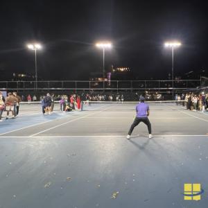 Tennis player in ready position at the baseline of a night-lit court, with three LED sports light poles overhead and spectators along the sidelines.