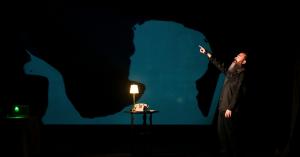 Comedian and filmmaker Jon Schnitzer with a long beard points to the left while a projection of a silhouette of his grandmother in a rocking chair is projected behind him. Something Spooky is a paranormal comedy show that will give you goosebumps.