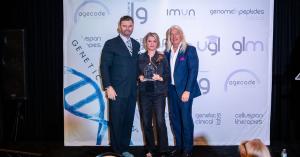 Three people pose on stage with a Genetic LifeSpan award in front of a backdrop showing AgeCode, IMUN, GLM and other Genetic LifeSpan brands