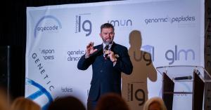 Adam Vincent Gilmer speaks into a microphone on stage in front of a backdrop showing AgeCode, LivinGene, IMUN and GLM logos