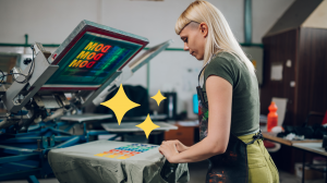 A woman with blonde hair works at a screen printing press in a workshop, pulling a gray shirt printed with colorful text off the platen, with decorative yellow sparkle graphics layered over the image and a screen showing repeated "MOD" text above her.