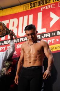 Isaac “Pitbull” Cruz stands shirtless beside his championship belt during fight week, with a PBC promotional backdrop behind him.