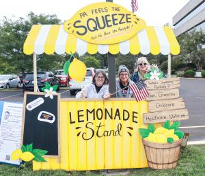 Three women standing at a lemonade stand; a sign on the lemonade stand reads "PA Feels the Squeeze."