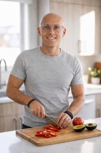 Fitness coach and author Sean Barker smiling in a bright kitchen while slicing red peppers, with a tomato and avocado on the cutting board