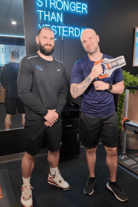 Race Director Brian Barao and MetaLab technician stand inside MetaLab by Confidia with the grip strength device featured in the “Can You Beat Brian?” challenge.