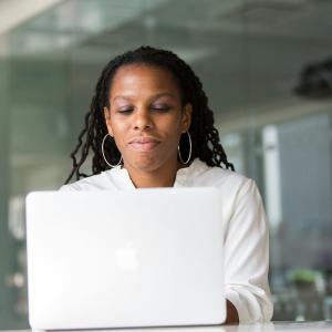 A woman with long braids sits at a laptop in a glass-walled office, focused on her screen
