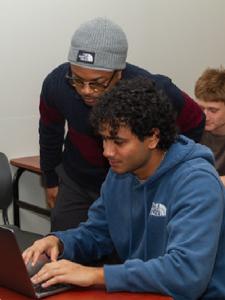Two people look at a laptop together. A student in a blue hoodie types while another person in a gray beanie and dark sweater leans in to watch the screen.