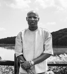 Black-and-white portrait of Joel Leggett in a light T-shirt standing with his arms crossed beside a lake, with wooded hills in the background.