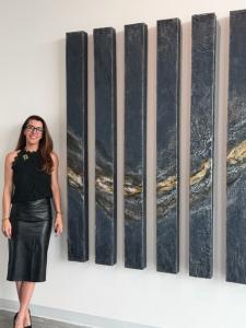 Artist Lauren Glad dressed in black poses beside her artwork made up of seven narrow panels mounted on a wall.