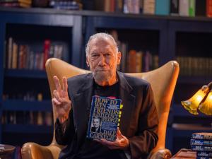 John Paul DeJoria poses in a high-backed leather library chair, holding a copy of "How to Win Friends and Influence People" by Dale Carnegie.