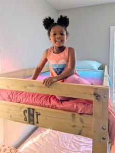 A smiling young girl with her hair in two puffs sits on the top of a wooden bunk bed. The SHP logo is branded into the front rail. The bed is made with bright pink, purple, and blue bedding, a light blue pillow, and a coral comforter, and a pink blanket c