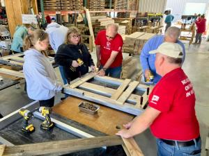 A group of Sleep in Heavenly Peace volunteers gathers around a workbench in a chapter workshop, assembling a wooden bed frame on a metal jig. A woman holds a yellow power drill while a volunteer in a red SHP shirt points to show her where to fasten the ne