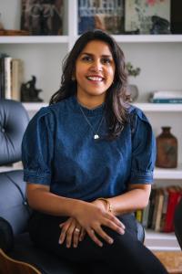 Aruna Mall, a woman with olive complexion, wearing a blue denim puffy sleeved top, gold jewelry, smiling and sitting in front of a shelf lined with books.
