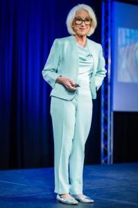 Judy K. Herman speaking on stage in a light blue suit while wearing a headset microphone and holding a presentation remote.