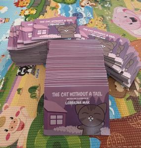 Stacks of The Cat Without a Tail picture books by Lerraina Mak, with purple covers and a smiling tabby cat, on a colorful mat.