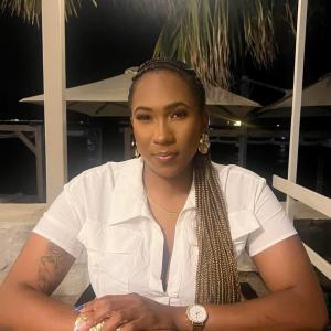 A woman with long braids resting over her shoulder, wearing a white short-sleeved button-up shirt, gold earrings, and a watch. She is sitting at an outdoor table at night, looking directly at the camera, with a rose tattoo visible on her right forearm. Th